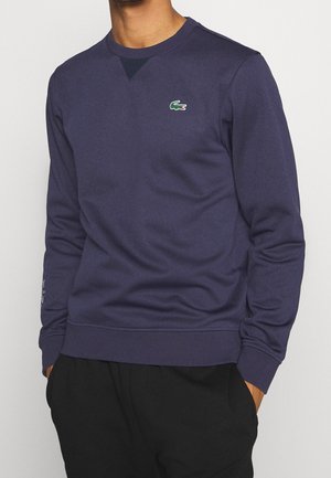 Man draagt een marineblauwe sweatshirt met lange mouwen en een klein groen krokodillenlogo op de linkerborst, handen in de zakken van zwarte broek.