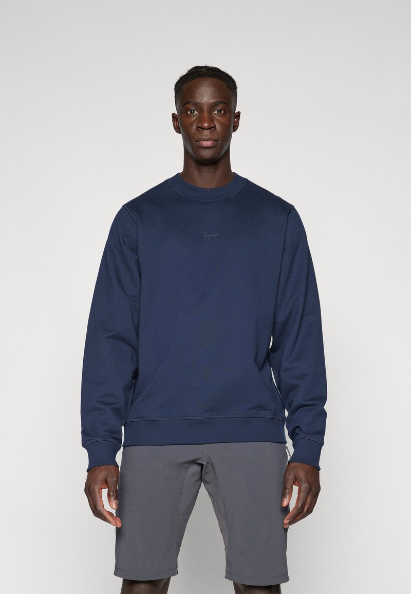 Mørkeblå sweatshirt laget av bomullsmiks med rund hals, ribbede mansjetter og en liten logo på brystet. Brukt med grå shorts.