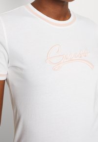 Person som bär en kortärmad vit t-shirt med ljusrosa kant och broderad "Guess"-logotyp på bröstet, beskuren vid nacke och överkropp.