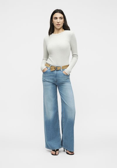 Νεαρή γυναίκα με μακριά σκούρα μαλλιά, που φοράει ανοιχτογκρι μακρυμάνικη μπλούζα, φαρδιά ξεβαμμένα μπλε τζιν, ταμπά ζώνη και μαύρα σανδάλια με τακούνια.