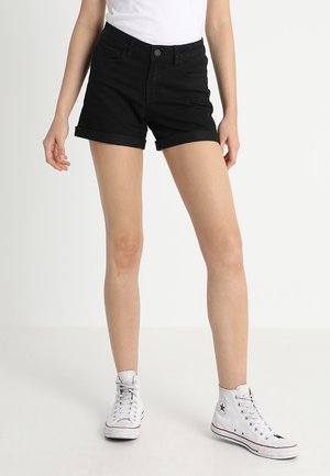 Person iført sorte shorts med opbukket kant, hvid t-shirt og hvide høje sneakers, står på en lys baggrund.
