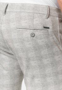 Pantaloni în carouri gri deschis cu o textură netedă, având un buzunar posterior și detalii cu buton. Modelul subtil adaugă un interes vizual.