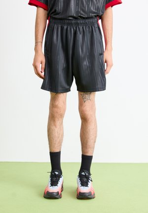 Mand i sorte Nike shorts med diskrete logoer, sorte sokker og atletik sko med hvid-til-rød gradient, stående på grøn gulv mod en hvid væg.