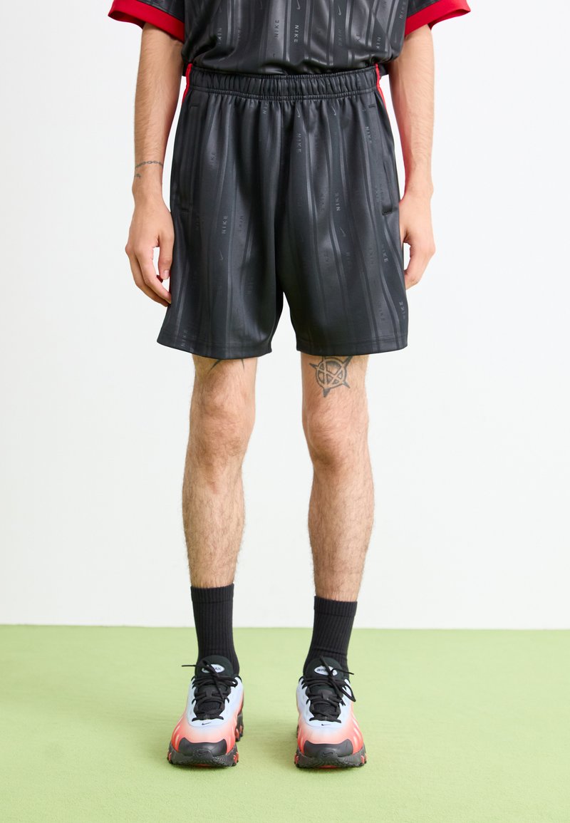 Uomo che indossa pantaloni corti neri Nike con loghi sottili, calze nere e scarpe da ginnastica con gradiente bianco-rosso, in piedi su un pavimento verde contro un muro bianco.