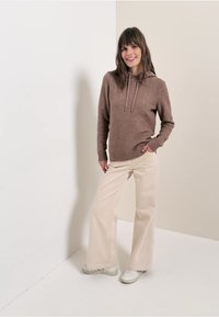 Brauner Hoodie mit Kordelzügen, kombiniert mit hellbeigen weiten Hosen. Modell trägt weiße Sneakers und steht vor einem neutralen Hintergrund.