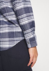 Personne portant une chemise à carreaux bleue et blanche à manches longues avec des boutons aux poignets et un pantalon sombre, main détendue sur le côté.