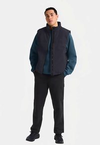 Zwart gewatteerd vest over een blauw sweatshirt, gecombineerd met zwarte cargobroeken en zwarte sneakers. Voorzien van een ritssluiting en een hoog kraagontwerp.