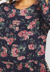 Kukkaismekko, jossa on laivastonsininen tausta ja vaaleanpunaiset ja vihreät kukat, pyöreä kaula-aukko ja laskostettu vyötärön yksityiskohta. Pehmeä kangaspinta.