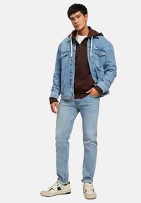 Jasnoniebieska kurtka dżinsowa o postarzanym wyglądzie, noszona na brązowej bluzie z kapturem, zestawiona z jasnoniebieskimi dżinsami i białymi sneakerami.
