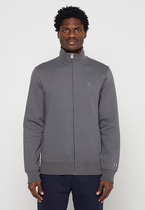 Hombre con sudadera gris con cremallera y cuello alto y pantalones azul marino, de pie frente a un fondo claro y simple.