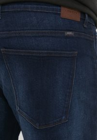 Vaqueros de mezclilla azul oscuro con una textura suave, que presentan una etiqueta de cuero y costuras en contraste. El bolsillo trasero incluye una etiqueta de la marca.