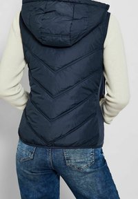 Námořnická pufovaná vesta má chevronové prošívání, kapuci a zipové zapínání, nošená přes svetřík světlé barvy a džíny.