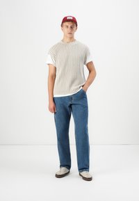 Top sans manches en tricot à côtes beige, porté sur un T-shirt blanc, associé à un jean droit bleu et à des baskets de couleur claire, le tout complété par une casquette rouge.