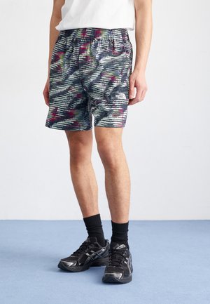 Mand iført flerfarvede kamuflage-mønstrede North Face-shorts, sorte træningssko, sorte sokker og en hvid skjorte, stående på blå gulv.