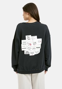 Frau in einem schwarzen Sweatshirt mit verschiedenen weißen Notizen auf dem Rücken, die motivierende Zitate und eine Einkaufsliste zeigen.