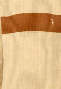 Sweter wełniany w kolorze kremowym z brązowym poziomym paskiem. Posiada strukturalne ściągacze oraz małe haftowane logo w dolnym rogu.