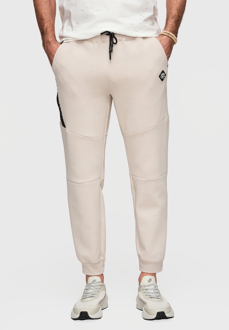 Homme portant un pantalon de jogging beige avec cordon noir, détail de poche latérale et chaussures de sport beiges, mains dans les poches devant un fond clair.