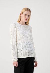 White long-sleeve blouse with eyelet patterns, round neck, and gathered shoulder detail. Paired with black trousers. Texture is airy and lightweight.