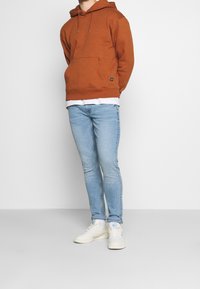 Osoba stojící s rukama za zády, oblečená v rezavě oranžové mikině s kapucí, světle modrých úzkých džínách a bílých teniskách s modrým logem.