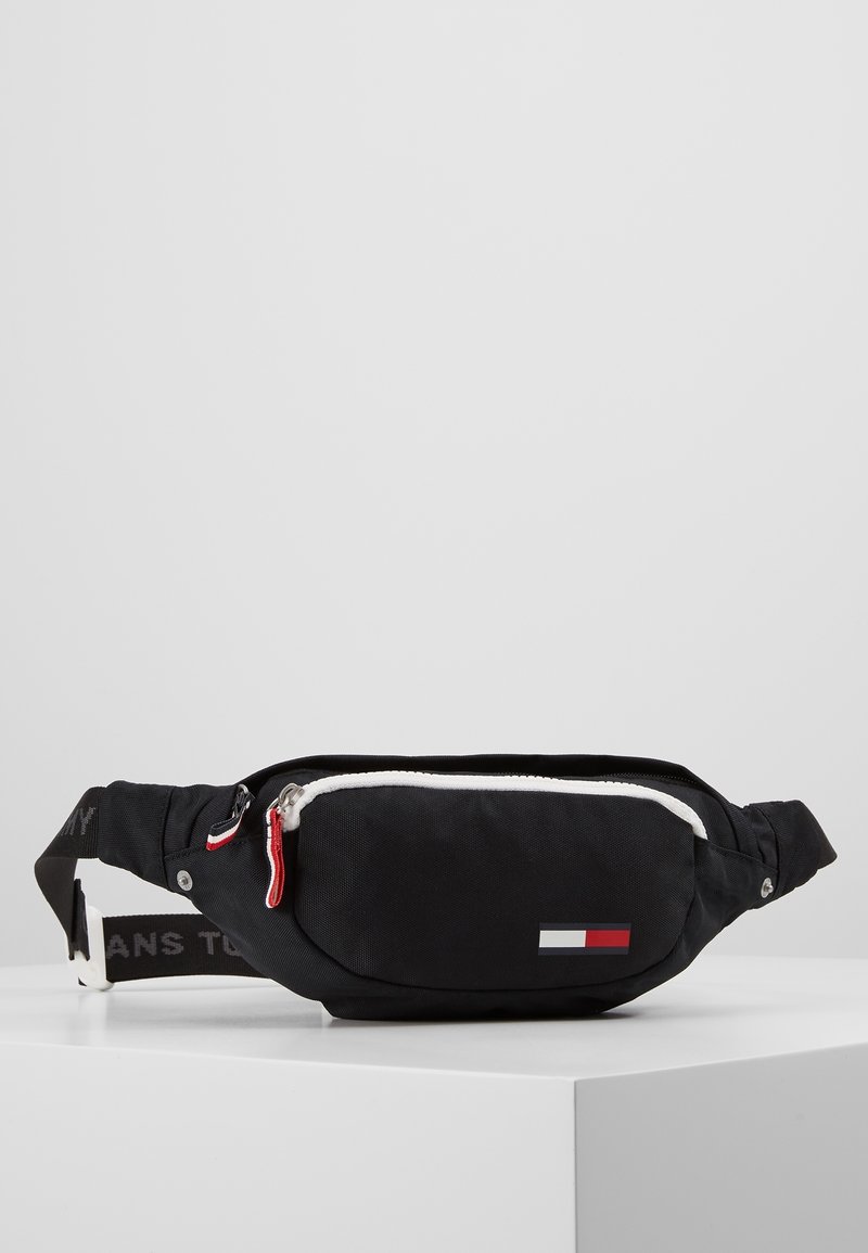 Sac banane en tissu noir avec fermeture éclair blanche et écusson logo rouge, blanc et marine, sangle ajustable avec texte de marque et tirette de fermeture éclair rouge.