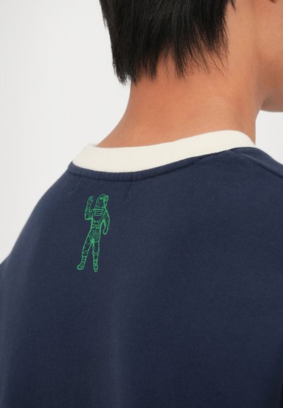Sweatshirt azul-marinho com colarinho branco em contraste, apresentando uma ilustração de um astronauta verde nas costas, perto da área do pescoço.
