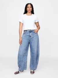 Hvit avkortet t-skjorte, vide lyseblå jeans med slitte detaljer, og grå joggesko med hvite såler. Uformelt antrekk mot en nøytral bakgrunn.