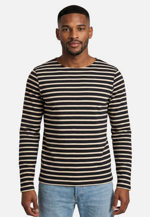 Modello maschile con capelli corti che indossa una maglietta a maniche lunghe a righe orizzontali nere e beige e jeans azzurri, in piedi di fronte a uno sfondo uniforme.