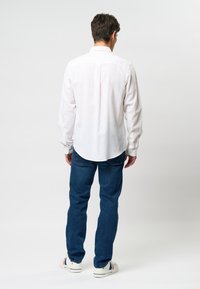 White long-sleeve shirt with a button-down collar, back pleat detail, paired with blue denim jeans and white sneakers with red accents.