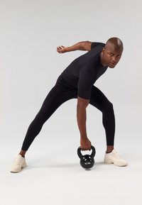 Top atlético preto e leggings, segurando um kettlebell preto de 10 kg. Ténis claros com detalhes texturizados. Foco na postura de exercício.