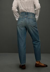 Džínsy svetlomodrej farby s vysokým pásom a voľným strihom, s zadnými vreckami a zatočeným lemom; nosené s tmavohnedými plochými topánkami.