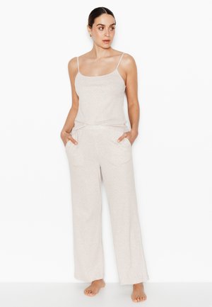 Donna in piedi a piedi nudi che indossa un top beige chiaro con spalline sottili e pantaloni larghi abbinati con le mani nelle tasche, che guarda di lato.