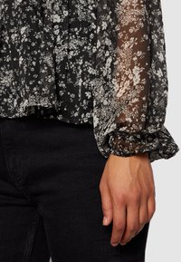 Czarna transparentna bluzka z białymi florystycznymi wzorami, plisowanym fasonem i zbieranym mankietem przy rękawie. Połączona z czarną dolną częścią.