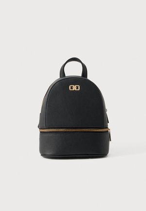 Sac à dos noir structuré, doté d’une poignée supérieure, d’un emblème doré à l’avant et de fermetures éclair dorées, sur un fond uni et clair.
