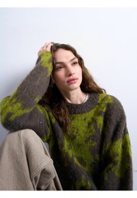 Jeune femme aux cheveux bruns ondulés portant un pull à motifs verts et gris, reposant sa tête sur sa main devant un fond blanc uni.
