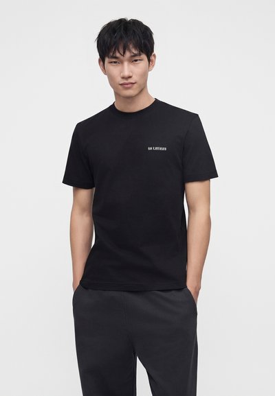 Νέος άνδρας που φοράει μαύρο T-shirt με μικρό λευκό κείμενο στο στήθος και σκούρο παντελόνι, στέκεται με τα χέρια στις τσέπες μπροστά σε απλό φόντο.