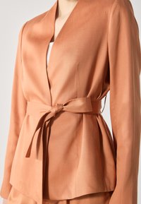 Peach-colored blazer made of smooth fabric, featuring a wrap design with a tied belt, pointed lapels, and subtle pleats.