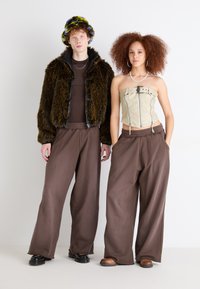 Braune weite Hose, cremefarbener strukturierten Oberteil mit Reißverschlüssen, schwarze flauschige Jacke und eine bunten Strickmütze. Flache Schuhe, neutraler Hintergrund.