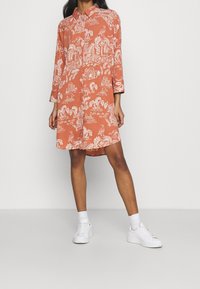 Sukienka w kolorze rdzy z długimi rękawami, kołnierzem i białym tropikalnym wzorem z palmami, budynkami i łodziami. Noszona z białymi trampkami.