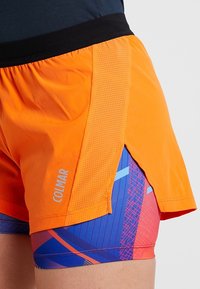 Oranga atletiska shorts med svart elastisk midjeband, med perforerad detalj och en färgglad geometrisk mönstring på det inre lagret.