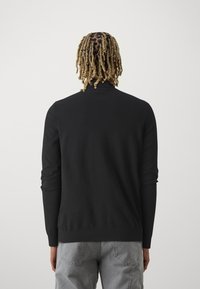 Czarny sweter z długim rękawem z wysokim dekoltem, o gładkiej fakturze i dopasowanym kroju, w połączeniu z szarymi spodniami typu cargo. Widok od tyłu.