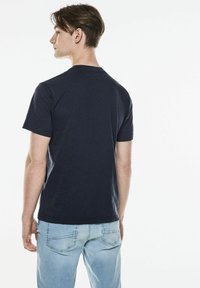 Tmavě modré bavlněné tričko s krátkými rukávy, kulatým výstřihem a volným střihem, v kombinaci se světle modrými džíny, z pohledu zezadu.