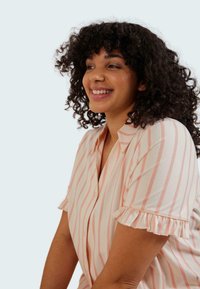 Chemise boutonnée rose pâle avec des rayures blanches et des manches volantes, fabriquée dans un tissu doux. Le modèle a des cheveux bouclés et sourit.