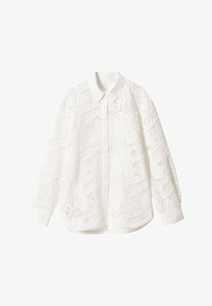 Mujer con camisa blanca de encaje con botones, vaqueros azules de pierna recta y sandalias blancas de tacón, de pie en un suelo liso con un fondo blanco.