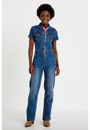Mujer sonriendo, vestida con un mono de denim azul de mangas cortas, cremallera frontal, pañuelo rosa en el cuello y botas marrones hasta el tobillo, de pie sobre fondo blanco.