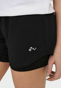 Gros plan d'une personne portant un short de sport noir à plusieurs couches avec un petit groupe de cinq cercles gris en logo sur la couche extérieure.