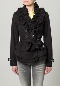 Svart jacka med volanger, dubbelknäppt stängning, peplumdesign och tygskärp. Har knappar och kontrasterande ärmslut.