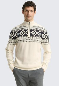 Strickpullover in Creme mit Reißverschlusskragen, der ein auffälliges schwarzes geometrisches Muster auf der Brust und kontrastierende blaue Akzente aufweist.