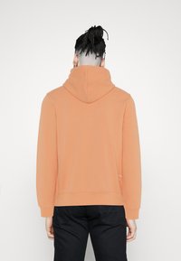 Hoodie orange avec une capuche à cordon, texture douce et poignets côtelés. Présente une marque minimaliste sur le côté inférieur, porté avec un pantalon noir.