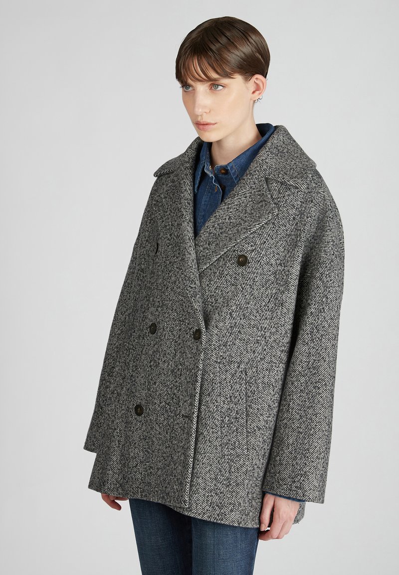 Cappotto in lana a spina di pesce grigio con ampio rever, design doppiopetto, grandi bottoni e vestibilità rilassata, indossato sopra una camicia di denim.