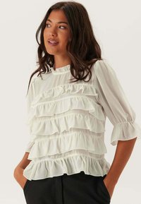 Blusa bianca con rouches e maniche lunghe, caratterizzata da strati a più livelli, un collo arricciato e una texture fluida. Pantaloni scuri sullo sfondo.